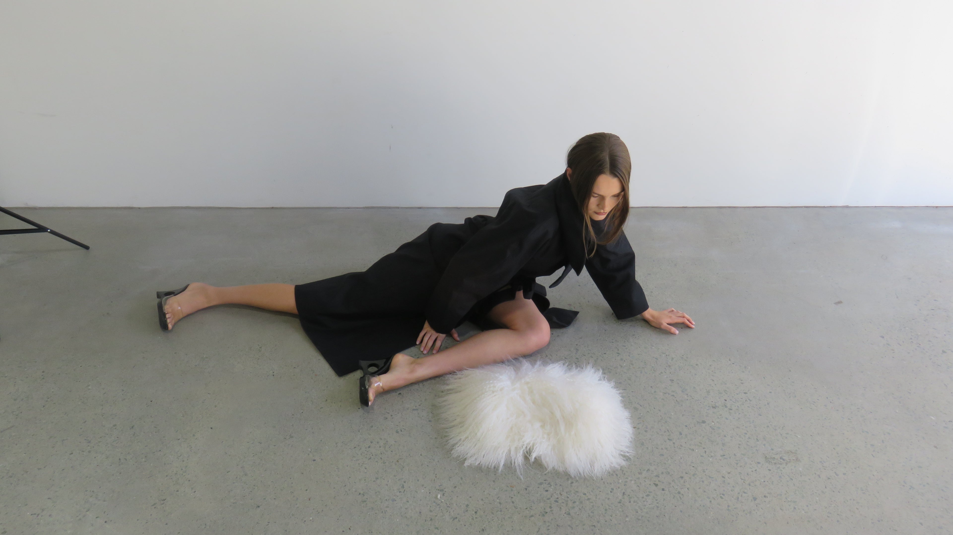 Woman in a black outfit sitting on a white fur rug against a plain background