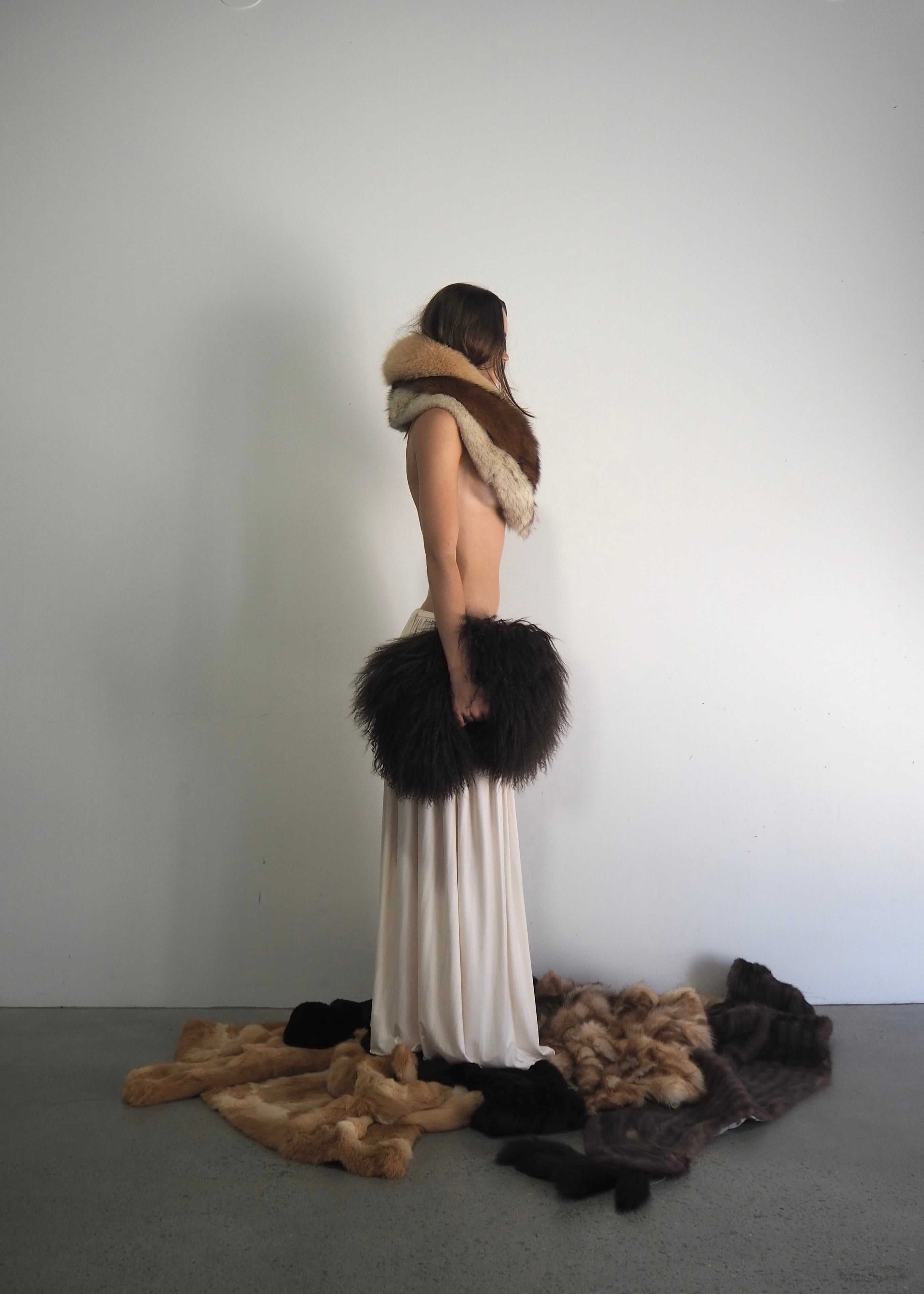 Person wearing a fur stole and holding a fur bag with various furs on the floor against a plain background